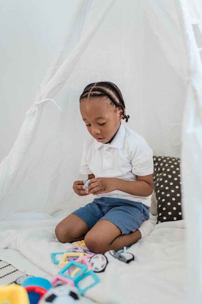 African American boy with cornrows playing with toys in a white teepee indoors.