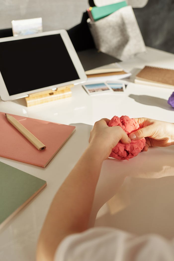 A child shaping pink clay on a cluttered desk with tablet and notebooks.